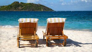 Deux chaises de plage en bois, sur lesquelles sont posées des serviettes à rayures, se trouvent sur une plage de sable, avec vue sur la mer et une petite île en arrière-plan.