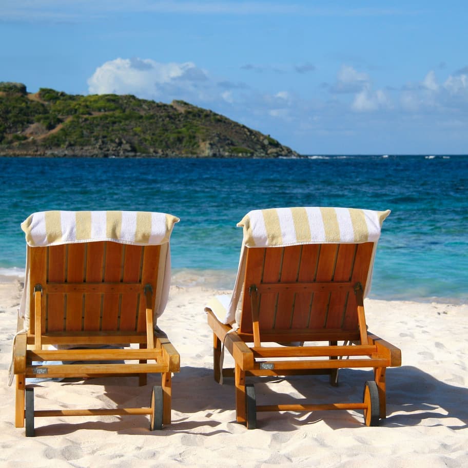 Two wooden beach chairs with striped towels on a sandy beach with a view of the sea and a small island in the background.