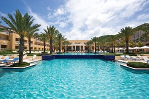 Ein großer Swimmingpool, umgeben von Palmen und Liegestühlen, mit einem Hotel im Hintergrund.