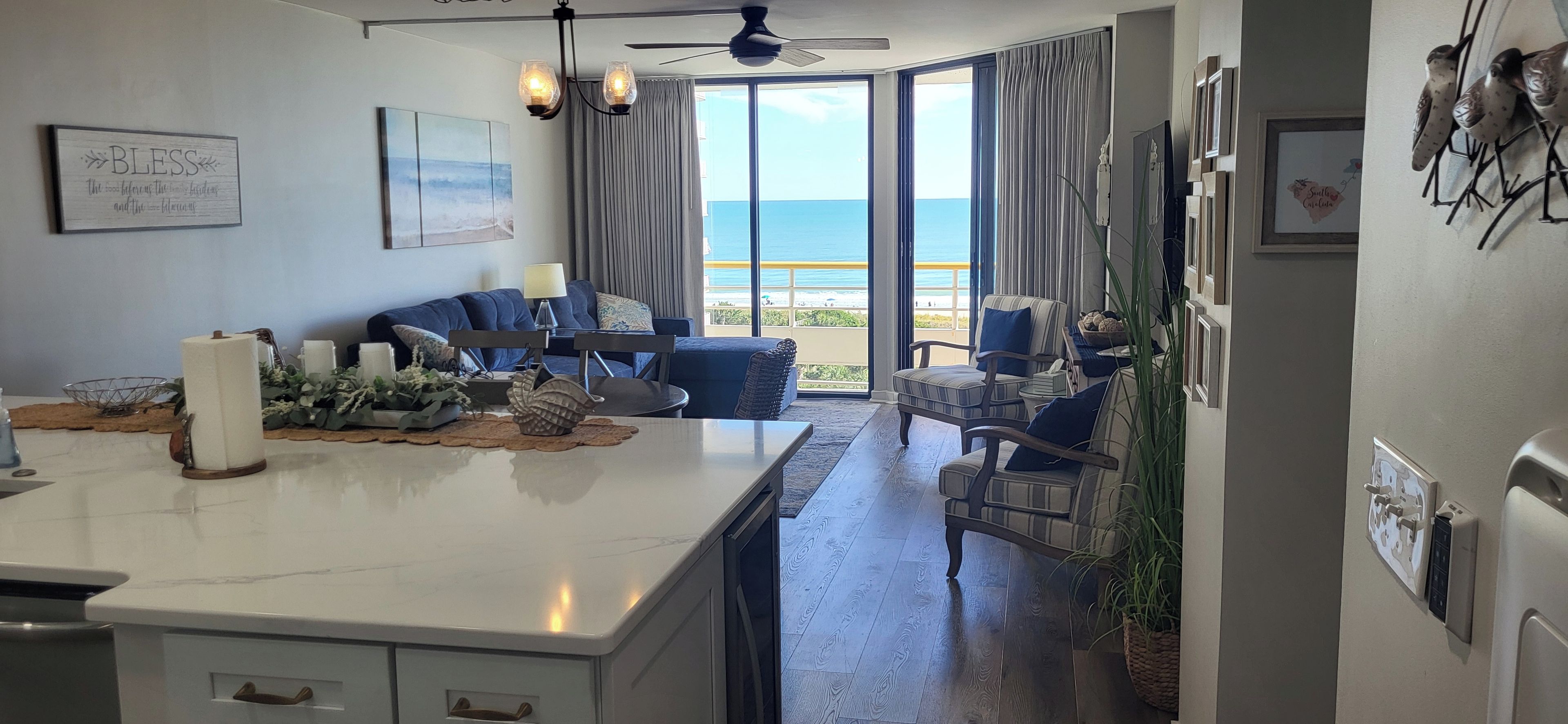 A modern kitchen with a large island, dining area with blue chairs, and a view of the ocean through large windows.