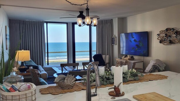 Un saló modern amb una finestra gran que dona a l'oceà, una zona de menjador amb una illa de marbre blanc i un televisor de pantalla plana penjat a la paret.