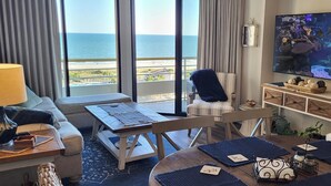 Habitació d'hotel amb una finestra gran que ofereix vistes a la platja i l'oceà, un televisor, una taula amb un ordinador portàtil i una zona d'estar còmoda amb un sofà i cadires.