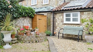 A stone house with a wooden door, a bench, and a garden with potted plants and pebbles. - Fantastic character cottage, brimming with charm and set in mature gardens. (Methwold, near Thetford)