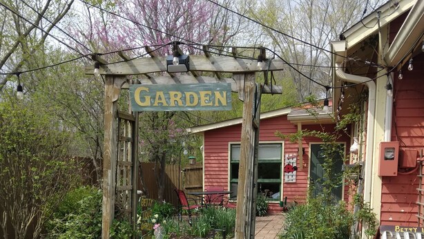 A garden entrance with a wooden archway labeled "GARDEN," a pathway leading to a red building, and a welcome sign. - Betty Lou's Garden Guest Suite (Nashville)