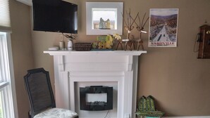 A living room with a fireplace, a television mounted on the wall, and a poster of Nashville, Indiana. - Betty Lou's Garden Guest Suite (Nashville)