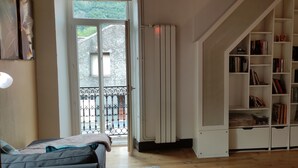 Une pièce avec un plancher en bois, un lit recouvert d’une couverture, une bibliothèque blanche et une vue sur un balcon doté d’une rambarde en métal.