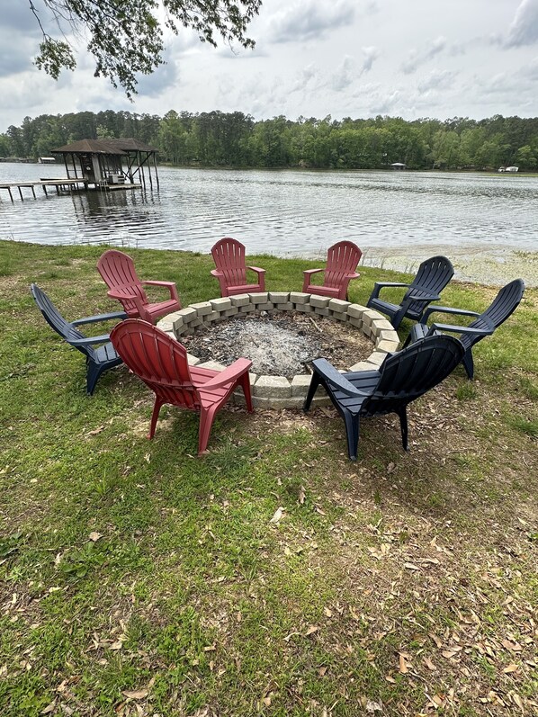 A circular fire pit with chairs around it, set on a grassy area by a lake.