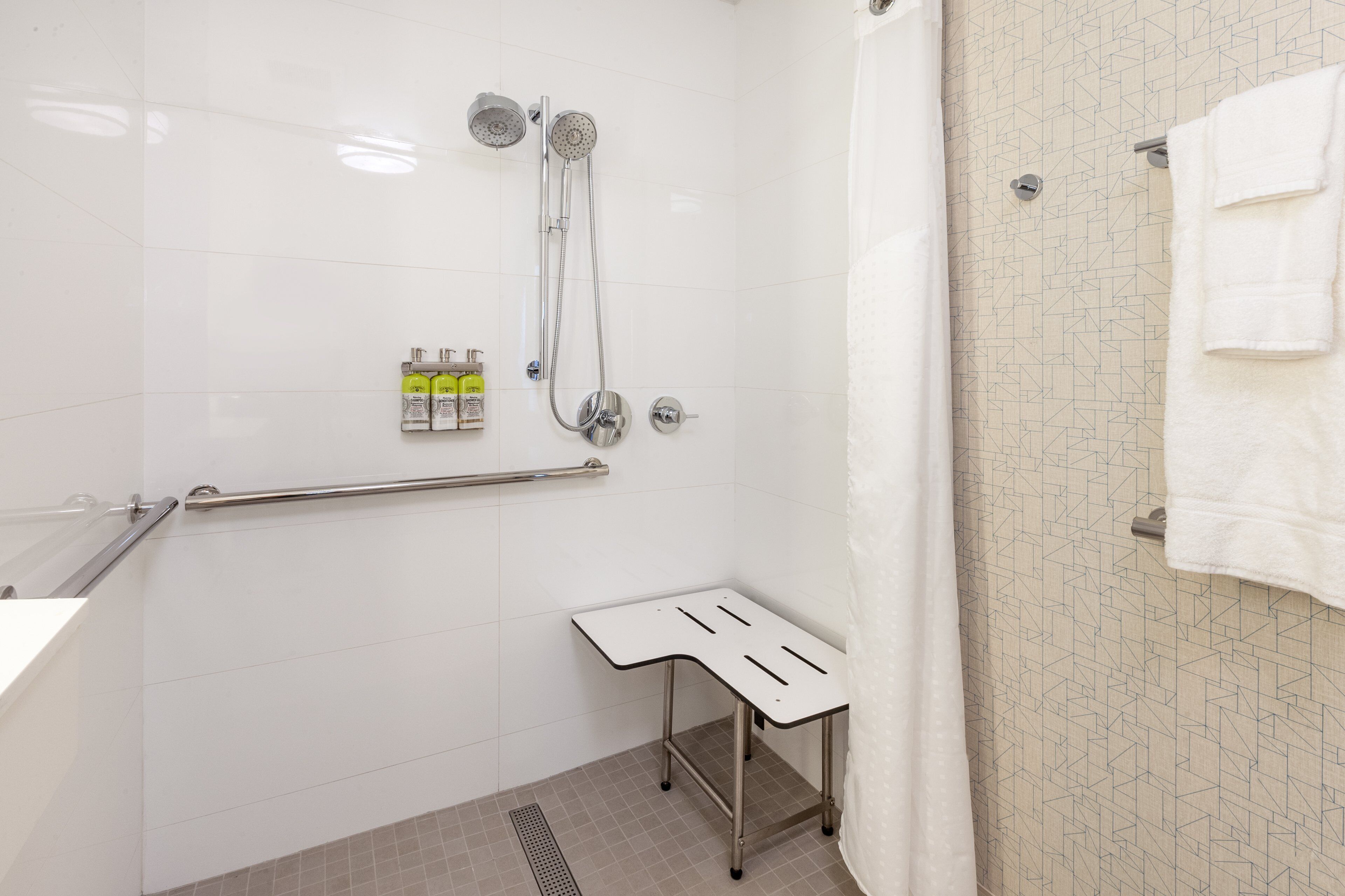 A bathroom with white tiles, a shower with a handheld showerhead, a grab bar, a small white bench, and a towel rack.