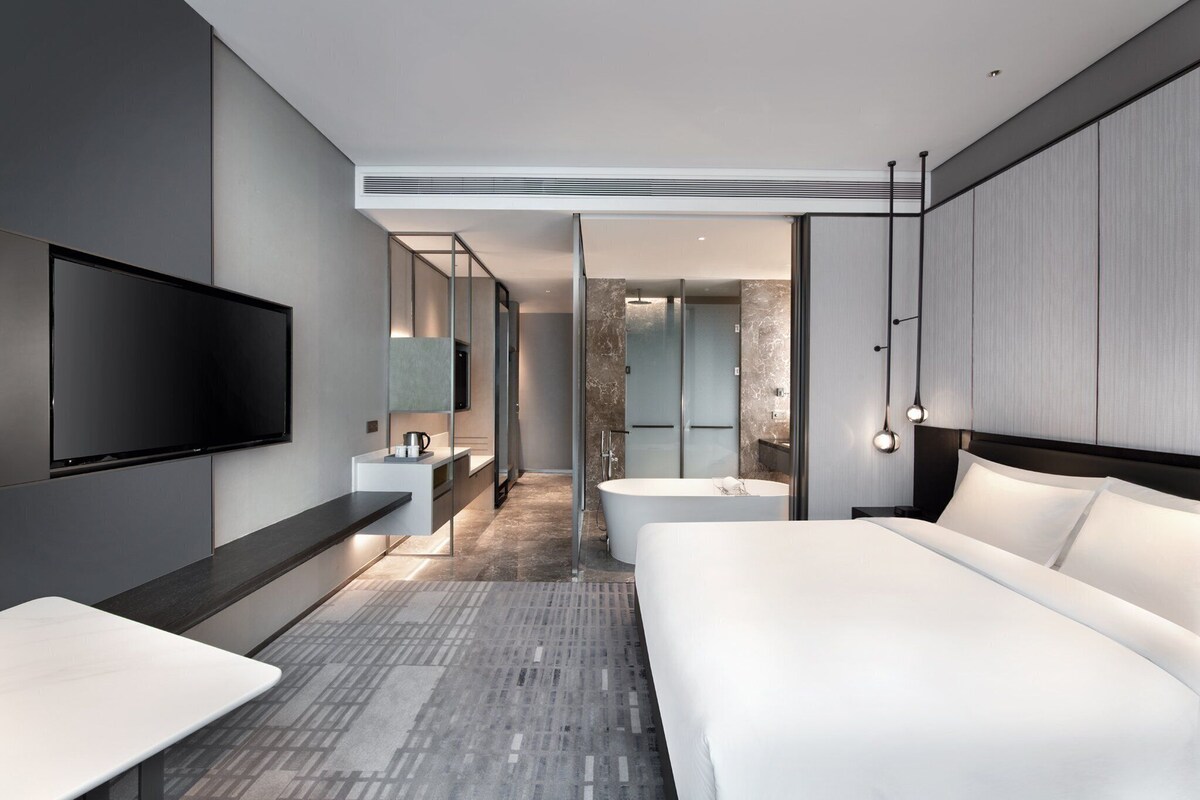 A modern hotel room with a large bed, a flat-screen TV, a desk, and a bathroom with a bathtub and shower.