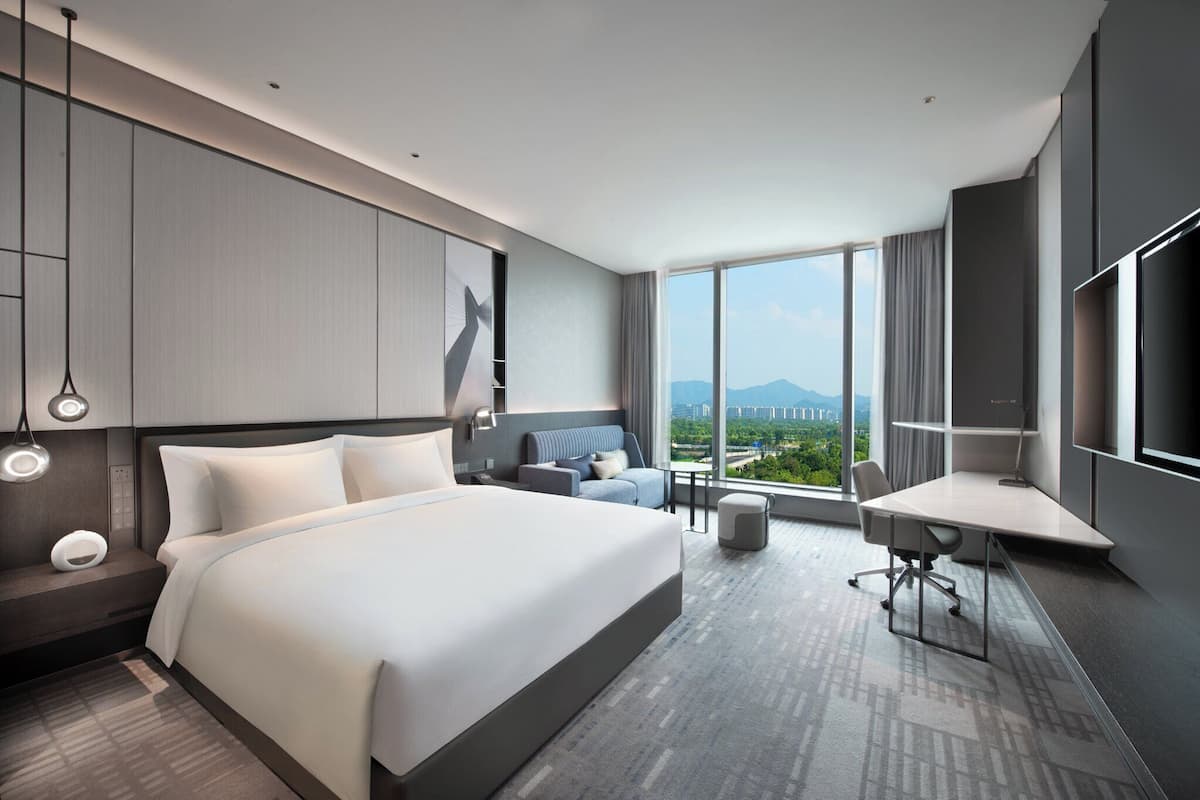 Quarto de hotel moderno com uma cama grande, uma escrivaninha e um sofá. Há uma janela ampla com vista para a cidade.