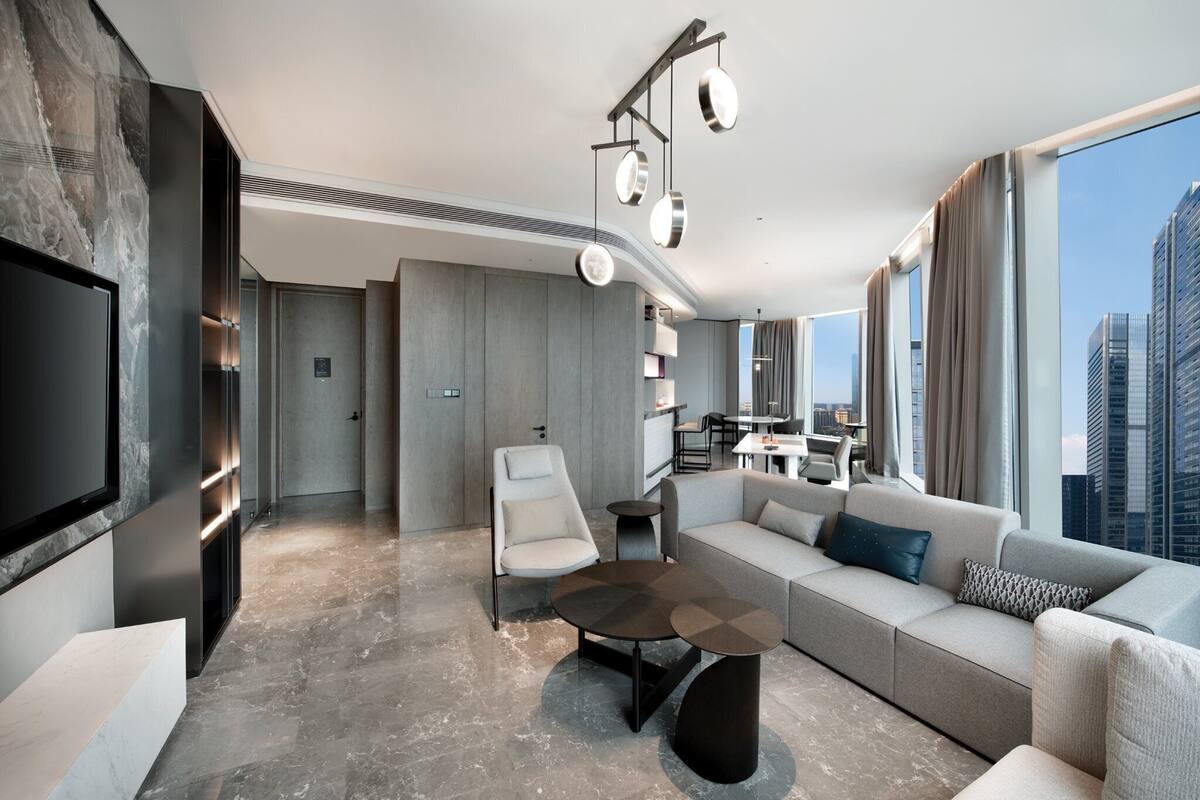 Uma sala de estar moderna com sofá, mesa de centro e TV. Há uma área de jantar com mesa e cadeiras. O quarto tem uma janela grande com vista para a cidade.