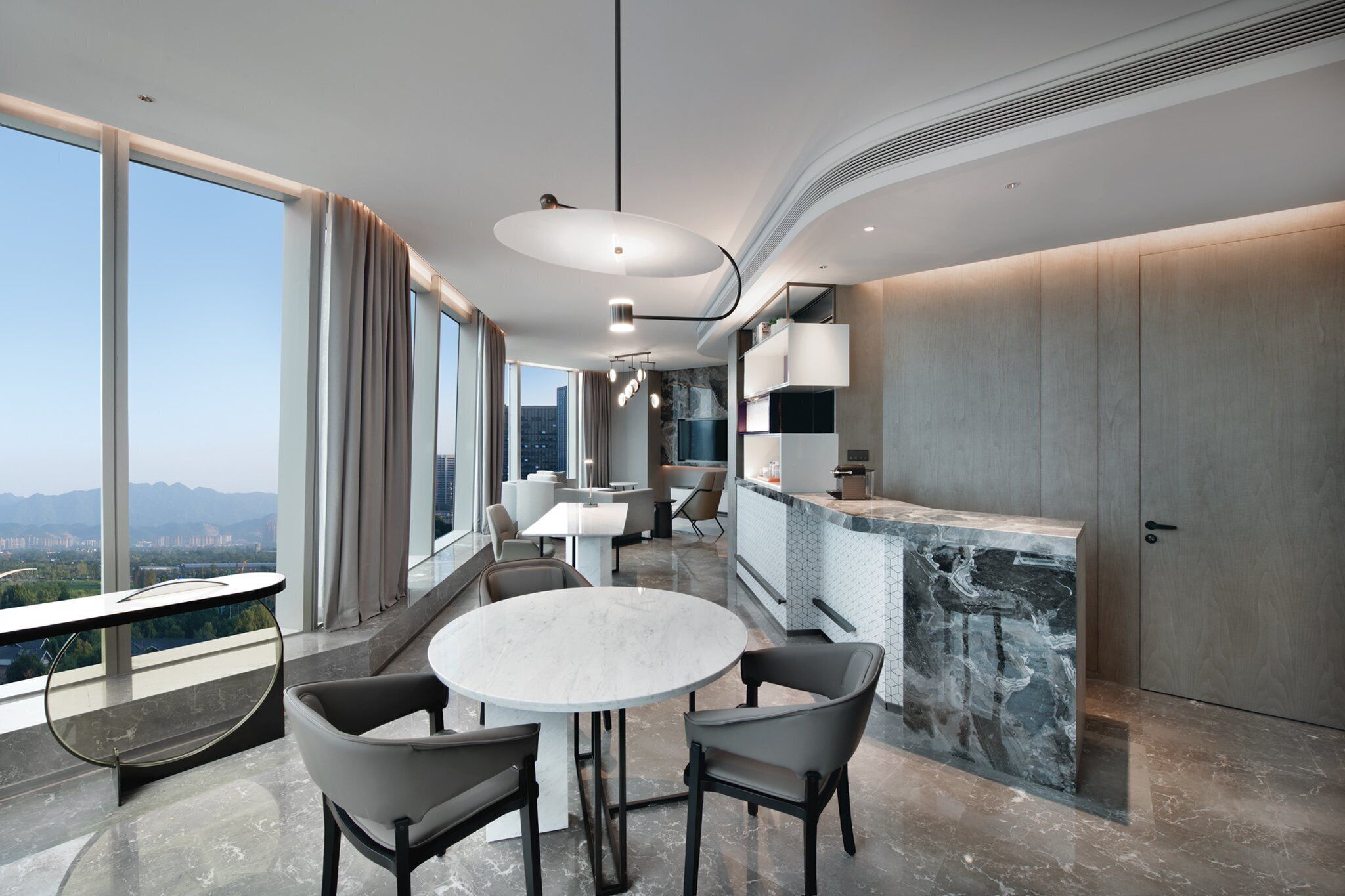 A modern dining area with a round table and chairs, a kitchen counter, and large windows offering a city view.