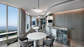 A modern dining area with a round table and chairs, a kitchen counter, and large windows offering a city view.