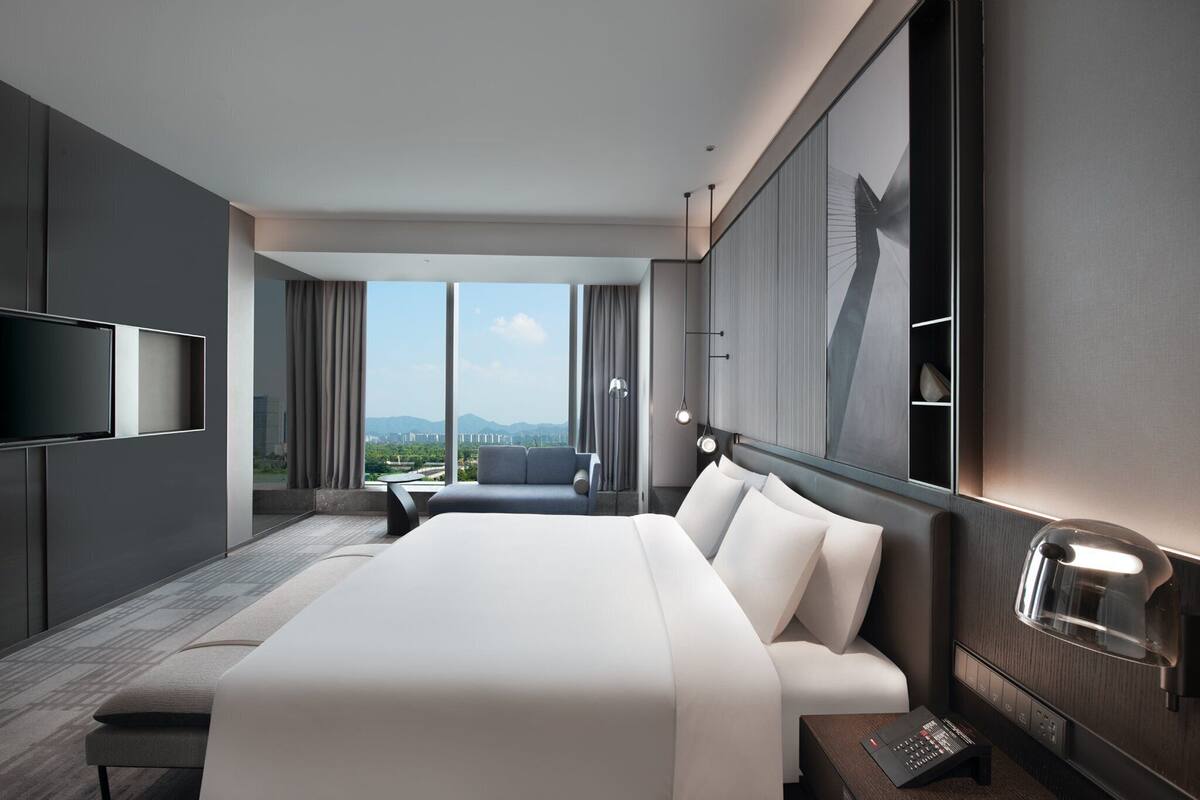 A modern hotel room with a large bed, a TV, a desk, and a view of the city.