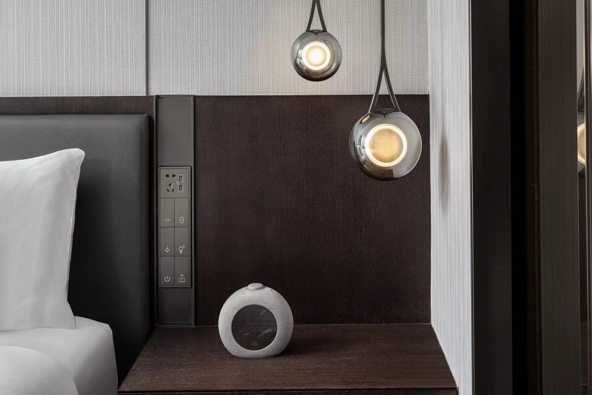 A modern hotel room with a bed, bedside table, and a control panel with a clock.