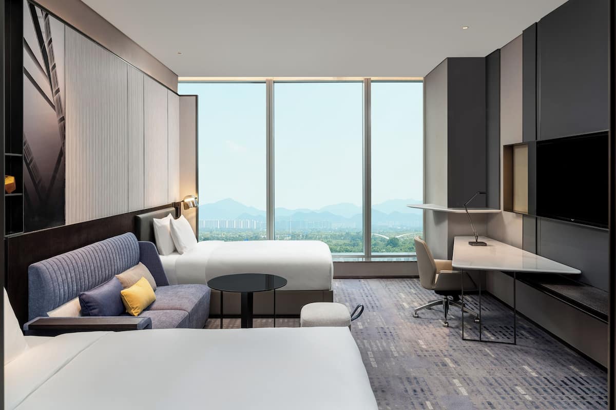Quarto de hotel moderno com uma cama grande, um sofá, uma escrivaninha e uma televisão. O quarto possui amplas janelas com vista para a cidade.