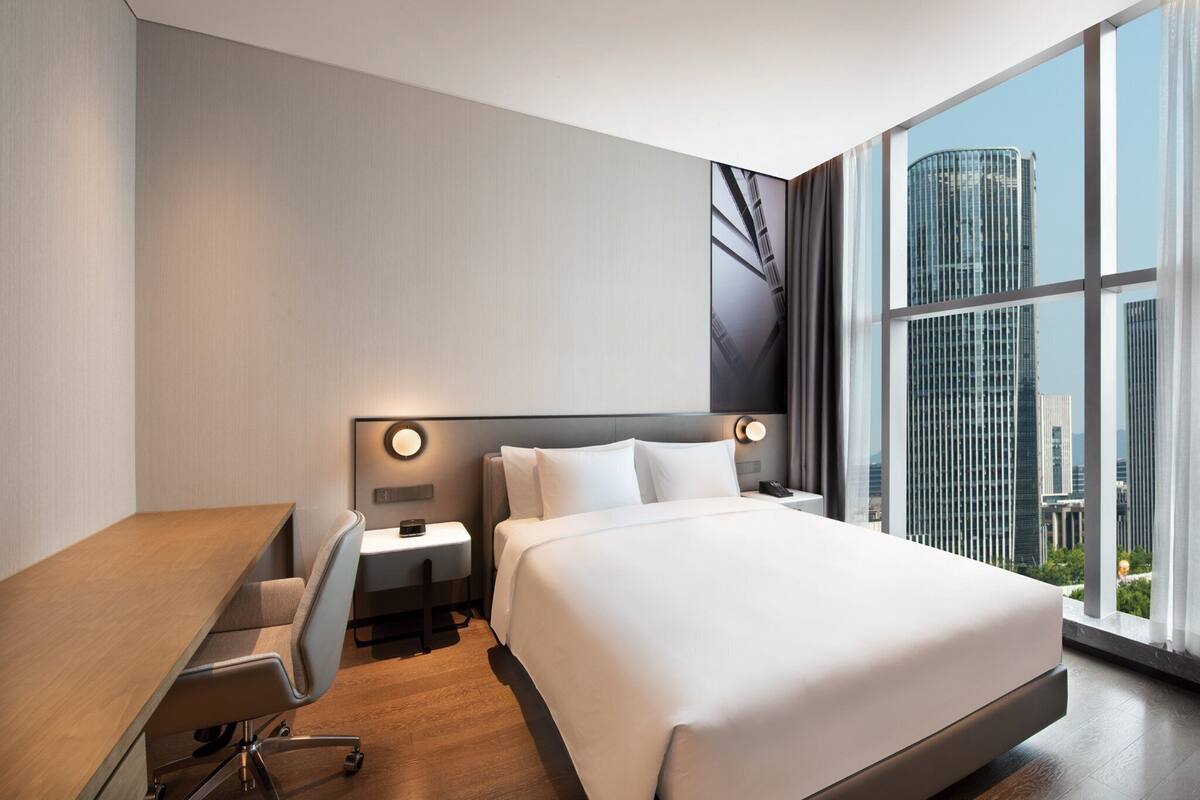 A modern hotel room with a large bed, a desk, and a view of a cityscape through large windows.