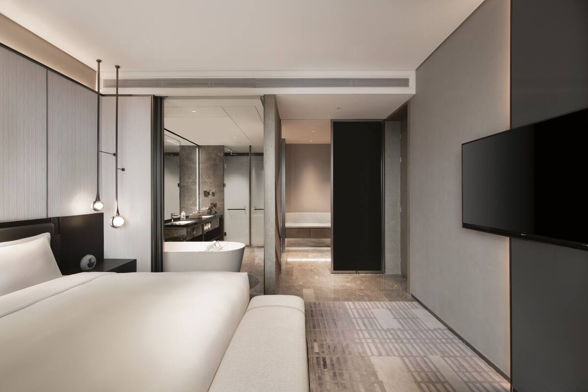 Quarto de hotel moderno com uma cama grande, televisão de tela plana, banheiro com banheira e um espelho amplo.