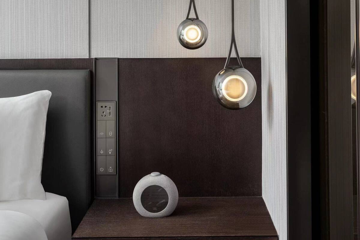 A modern hotel room with a bed, bedside table, and a control panel with a clock.