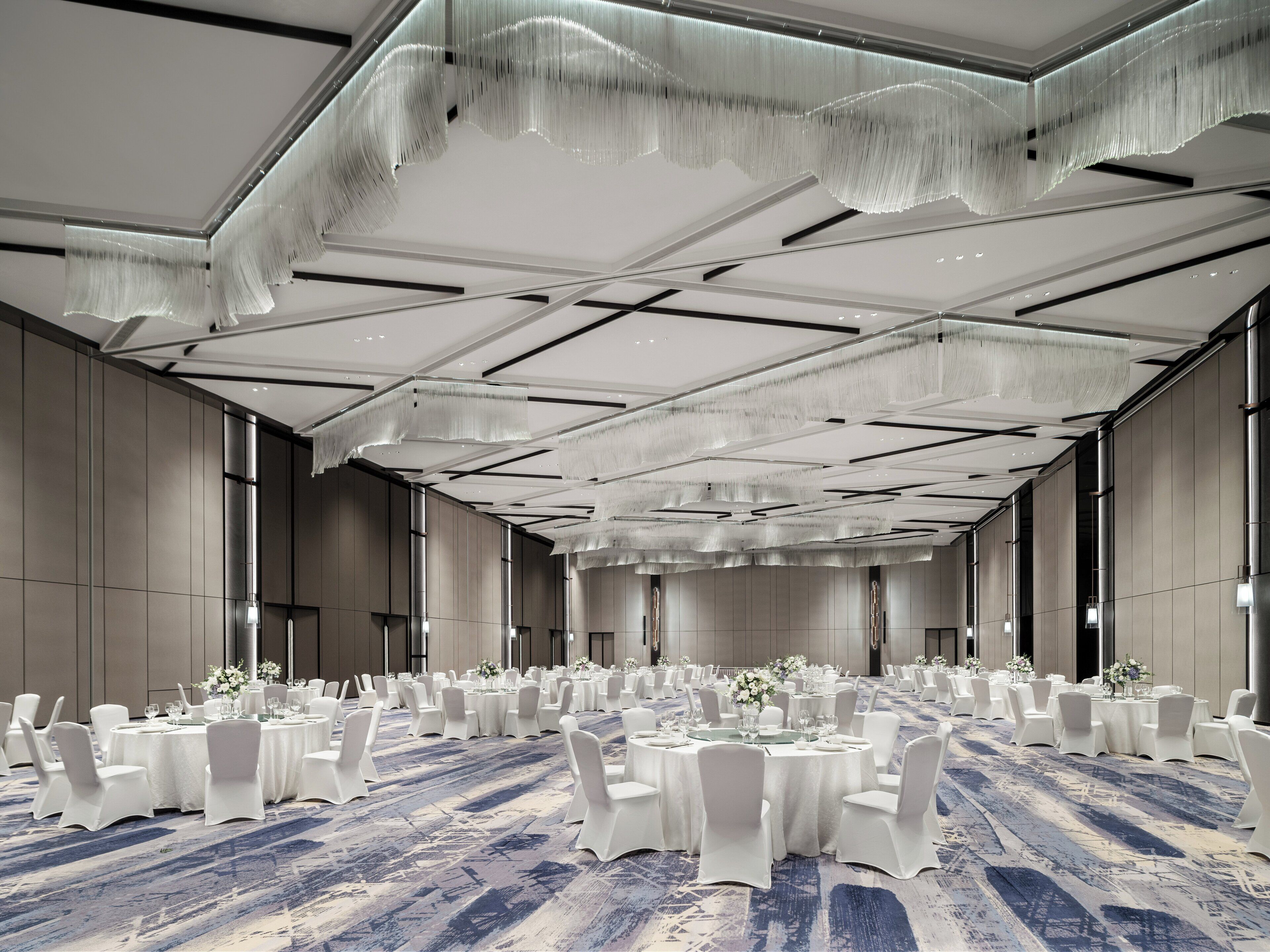 A spacious banquet hall with round tables and white chairs, a patterned carpet, and a modern ceiling design.