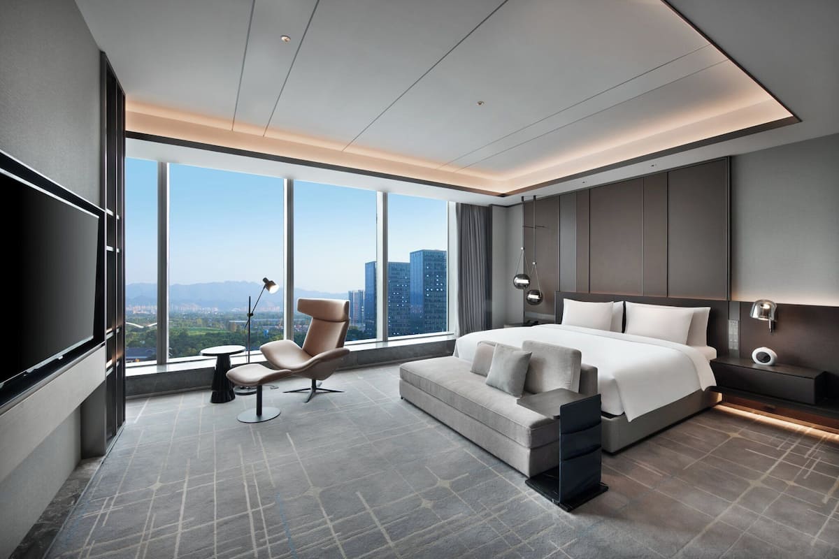 Quarto de hotel moderno com uma cama grande, área de estar, televisão e vista para a cidade.