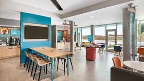 A modern restaurant with a large TV screen, wooden tables, and chairs. There is a counter with a coffee machine and a sign that reads "Fresh for you. All day. All night." The room has large windows and a view of the outside. - avid hotel Hays by IHG (Hays)