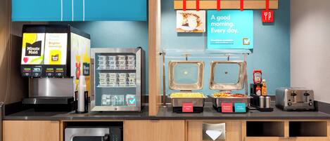 A breakfast area with a coffee machine, a juice dispenser, and a variety of food options including scrambled eggs, breakfast potatoes, and bacon.