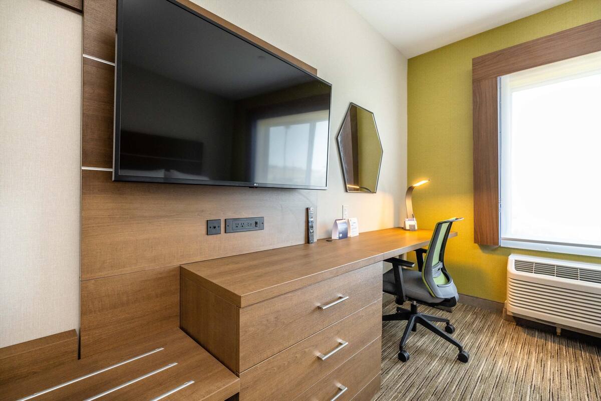 A hotel room with a wooden desk, a flat-screen TV, a chair, a bedside table, and a window with a view.