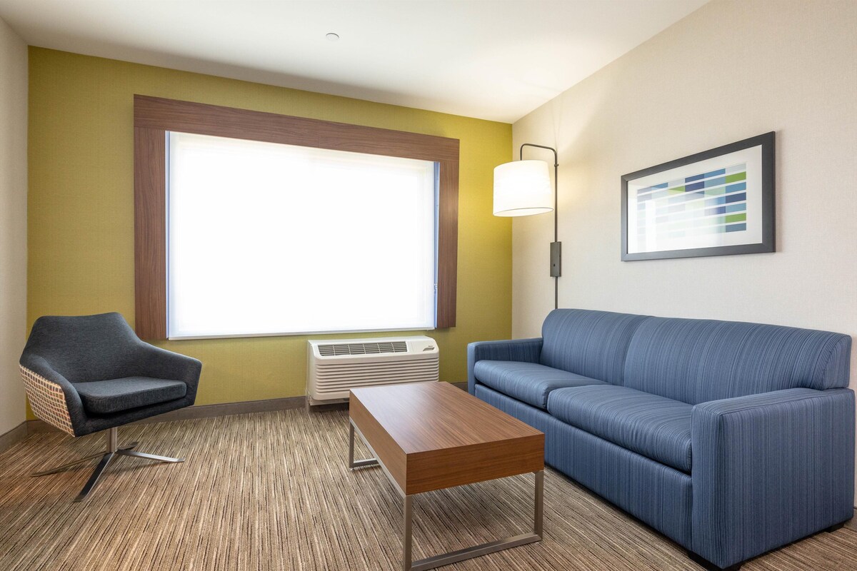 A hotel room with a blue sofa, a grey armchair, a wooden coffee table, a large window, an air conditioner, and a wall-mounted lamp.