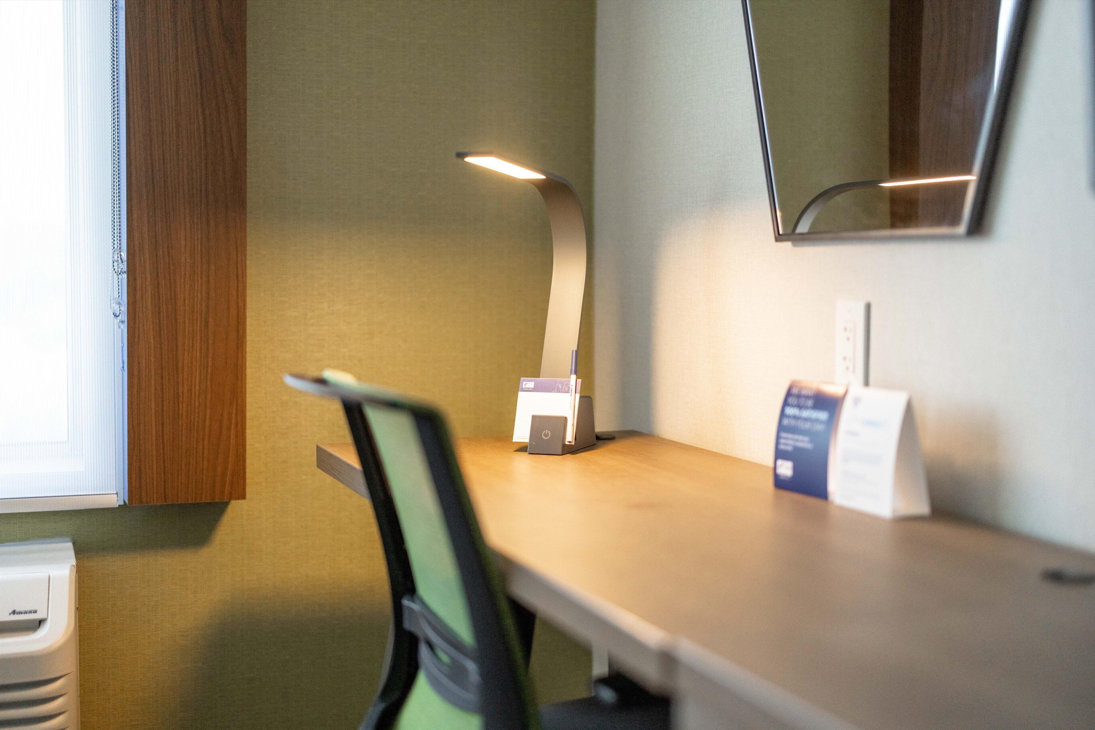 A hotel room with a wooden desk, a chair, a desk lamp, a small printer, and a mirror on the wall.
