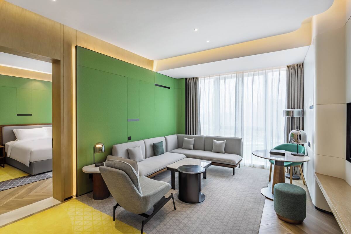 A modern hotel room with a sectional sofa, a single bed, a small round table, and a green accent wall.