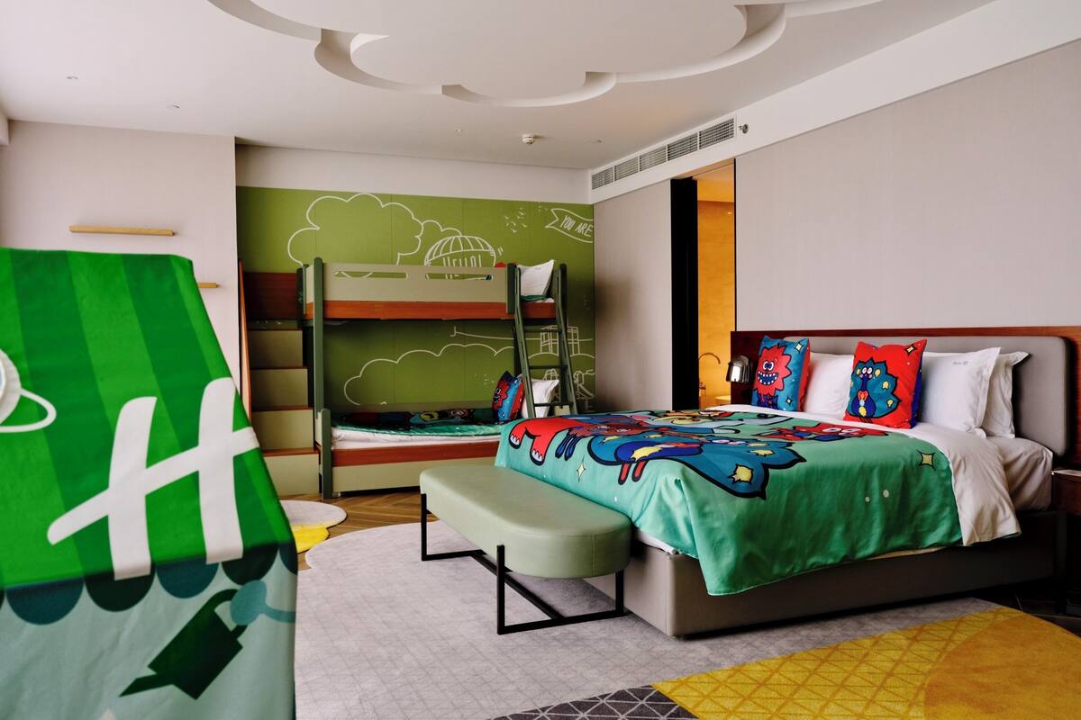 A hotel room with bunk beds, a bed with colorful bedding, a green wall with cloud drawings, and a white ceiling.