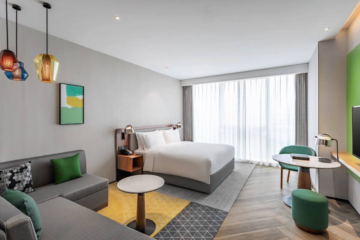 A modern hotel room with a large bed, a sofa, a small table, and a desk with a chair.