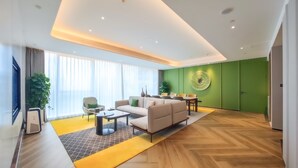 A modern living room with a sofa, coffee table, and a dining area with chairs. There is a large window with curtains, a green accent wall, and a ceiling with recessed lighting. - Holiday Inn Xi an Chanba by IHG (Xi'an)