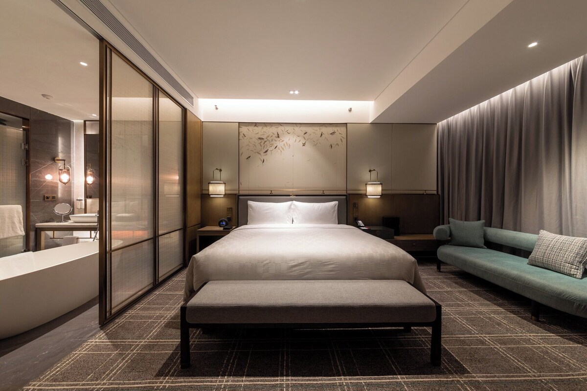 A modern hotel room with a large bed, a sofa, a bathroom with a bathtub, and a wall-mounted artwork.