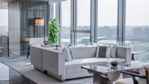 Ein modernes Wohnzimmer mit einer großen Couch, einem Couchtisch und Blick auf eine Stadtlandschaft durch große Fenster.