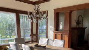 Een eetkamer met een houten tafel, vier stoelen, een kroonluchter en uitzicht op het groen door grote ramen.
