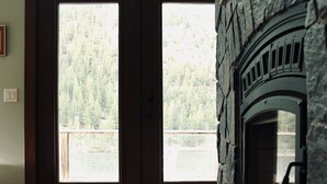 A room with a stone fireplace, wooden flooring, and a large window overlooking a forested area.