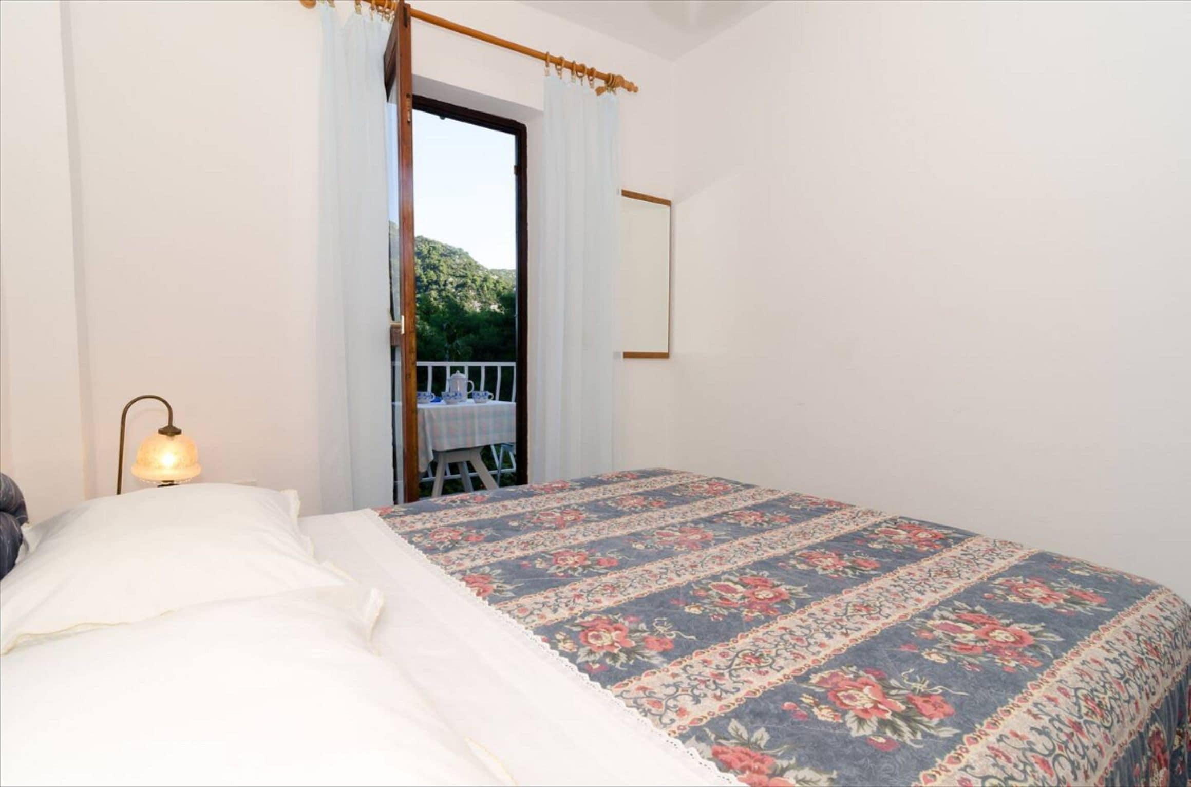 Un dormitorio con cama, mesita de noche, lámpara, ventana con cortinas y vistas a un balcón.