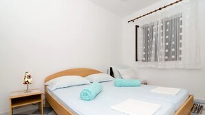 Ein ordentlich gemachtes Bett mit weißen Leinen und einem blauen Handtuch, ein kleiner Nachttisch mit einer Lampe und ein Fenster mit durchscheinenden Vorhängen.