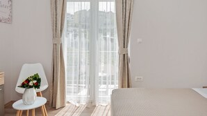 Ein Schlafzimmer mit Bett, einem Sessel, einem kleinen Tisch mit Blumenvase und einem Fenster mit Vorhängen.