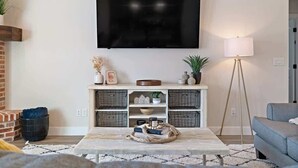 Ein Wohnzimmer mit an der Wand montiertem Fernseher, einer Couch, einem Couchtisch mit Dekoration, einer Stehlampe und einem Kamin.