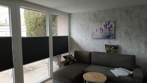 A modern living room with a grey sectional sofa, a round wooden side table, and a large window with blinds. - Ferienhaus in See Nähe! Ruhige Lage. Wlan, Bettwäsche und Handtücher Inkl (Klein Sien)