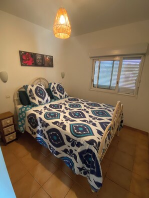 Une chambre à coucher comprenant un lit, une table de chevet, une fenêtre avec des stores et un luminaire suspendu.