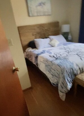A bedroom with a wooden headboard, a bed with a patterned bedspread, a nightstand with a lamp, and a door to the left. - Overlook Ninigret Pond and Block Island (Charlestown)