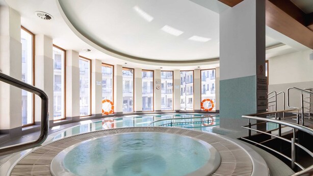 A large indoor swimming pool with a central hot tub, surrounded by glass walls and windows.