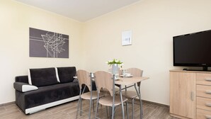 A modern living room with a sofa, dining table, chairs, a flat-screen TV, and artwork on the walls. - Forest Apartment with Balcony | 2.5 km from the beach (Wrzosowo)