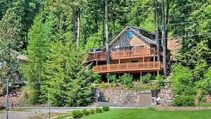 A two-story wooden cabin with a balcony, surrounded by dense forest and a body of water.