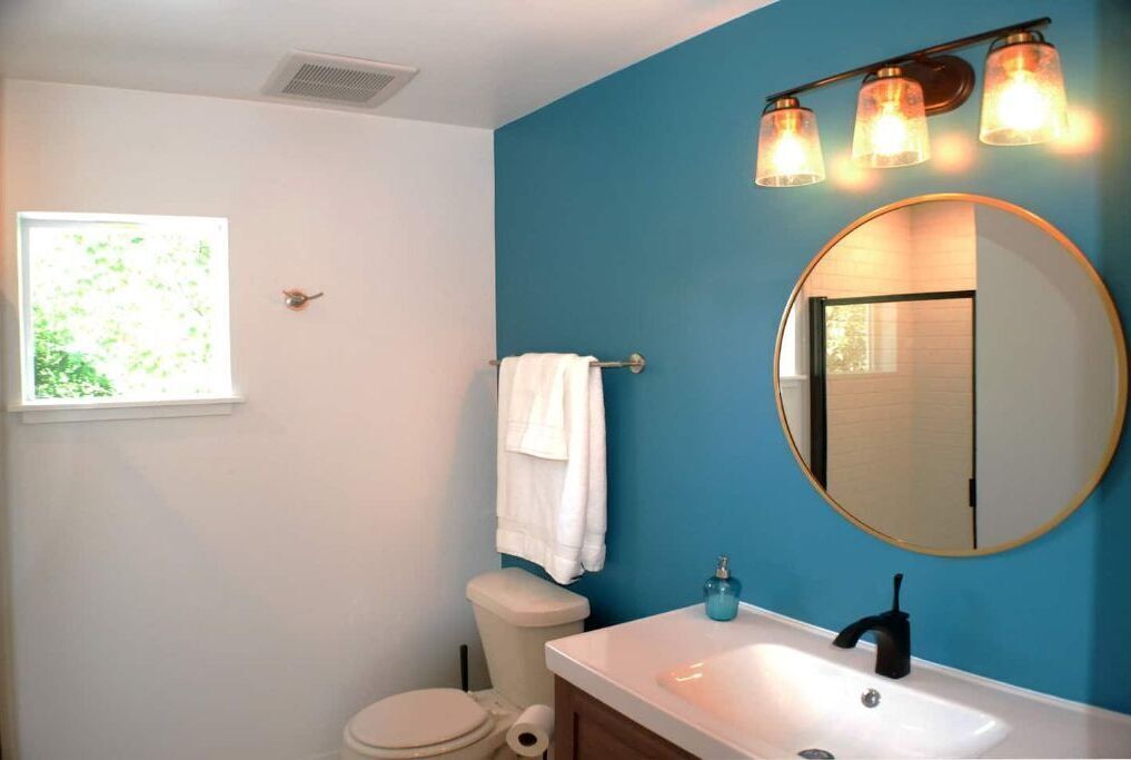 A bathroom with a white toilet, a white sink, a blue wall, a round mirror, and a window with daylight.