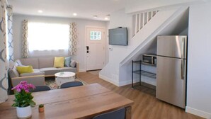 Un espacio habitable moderno con una mesa de comedor de madera, un conjunto de sofás, una escalera, una cocina con refrigerador y un televisor de pantalla plana montado en la pared.